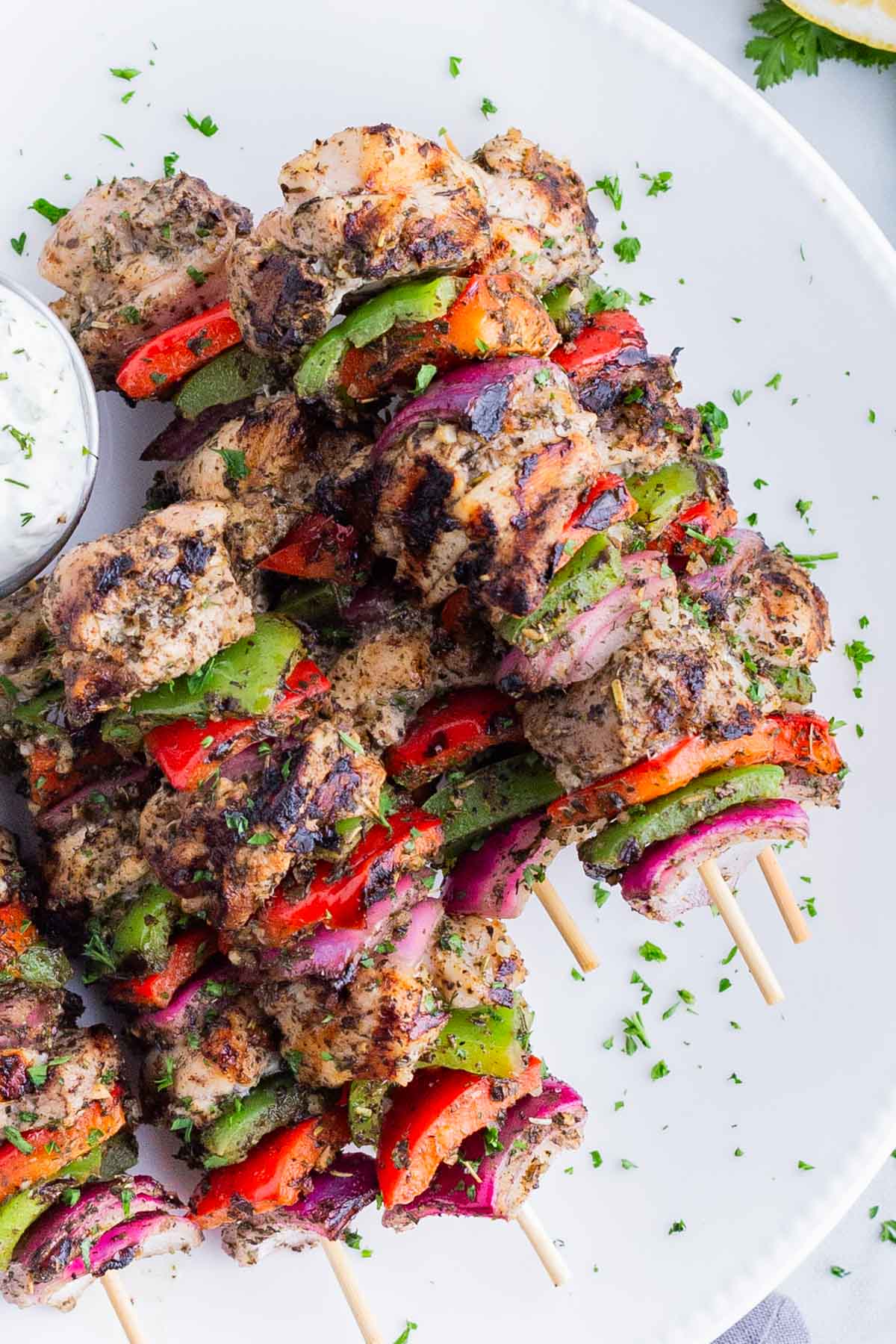 Grilled chicken kabobs with peppers and red onion are served on a white plate with Tzatziki sauce.