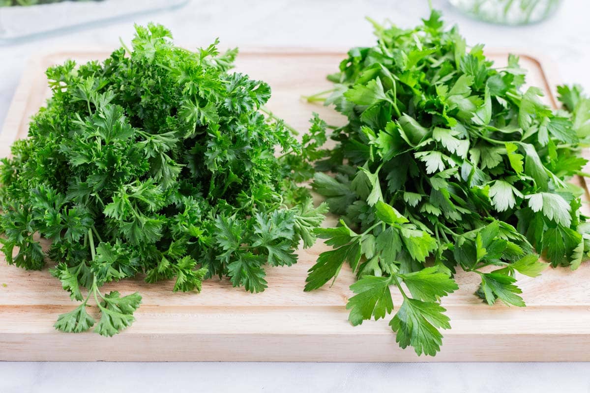 Flat leaf, or Italian parsley, and curly leaf parsley are laid on a cutting board.