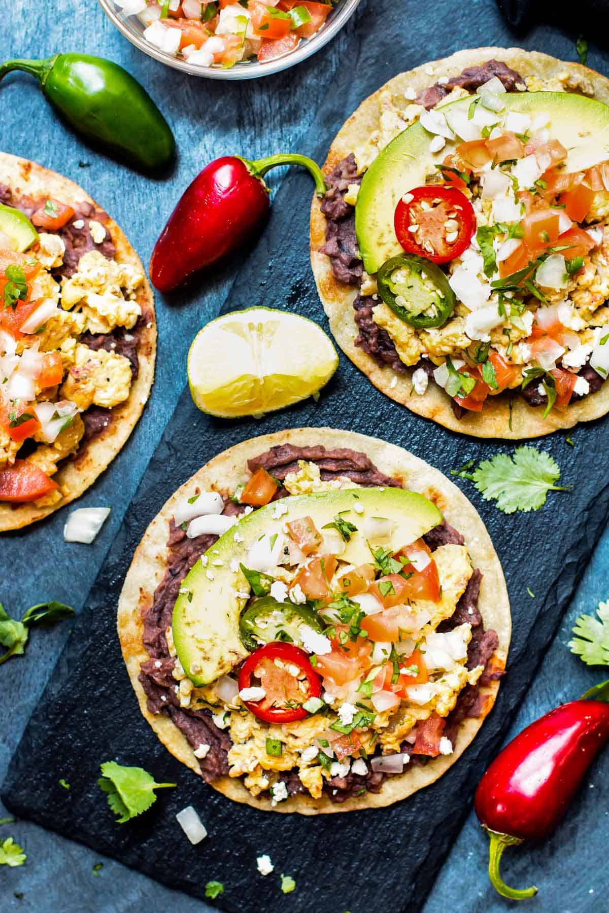 Three breakfast tostadas on a blue slab with peppers on the side.