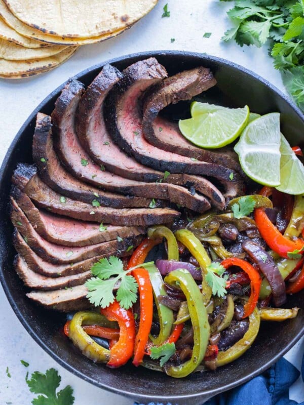 Beef flank steak fajitas are served in a cast iron skillet with fajita veggies and lime wedges.