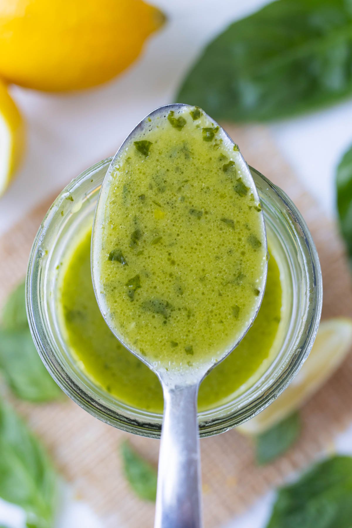 A spoon is holding a homemade lemon basil vinaigrette over a glass mason jar.