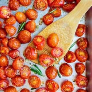 A spatula scoops up some roasted cherry tomatoes with herbs and garlic from a baking sheet.