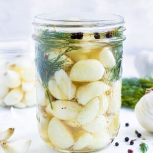 A jar of quick pickled garlic is on a table next to a head of garlic, peppercorns, and dill.