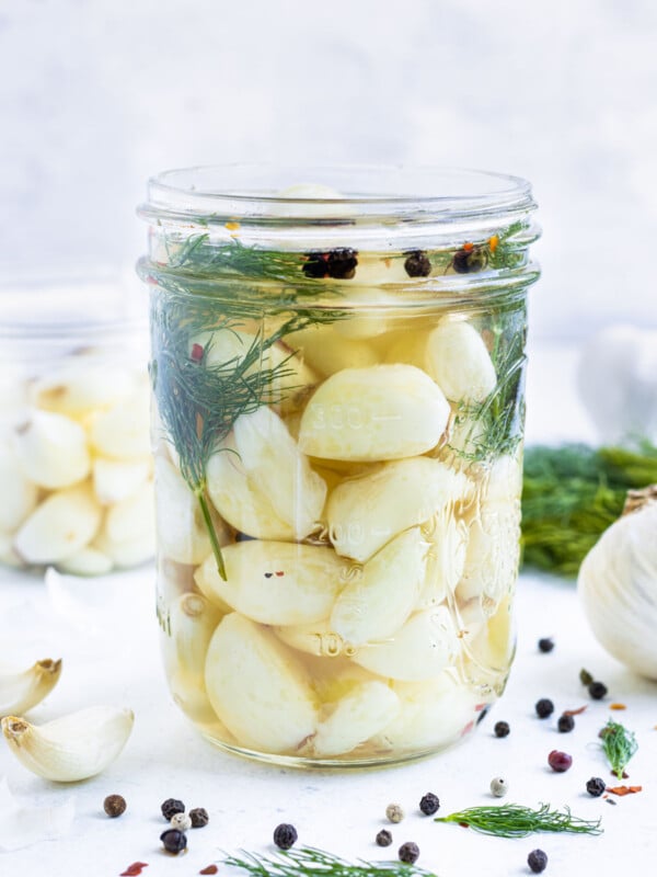 A jar of quick pickled garlic is on a table next to a head of garlic, peppercorns, and dill.