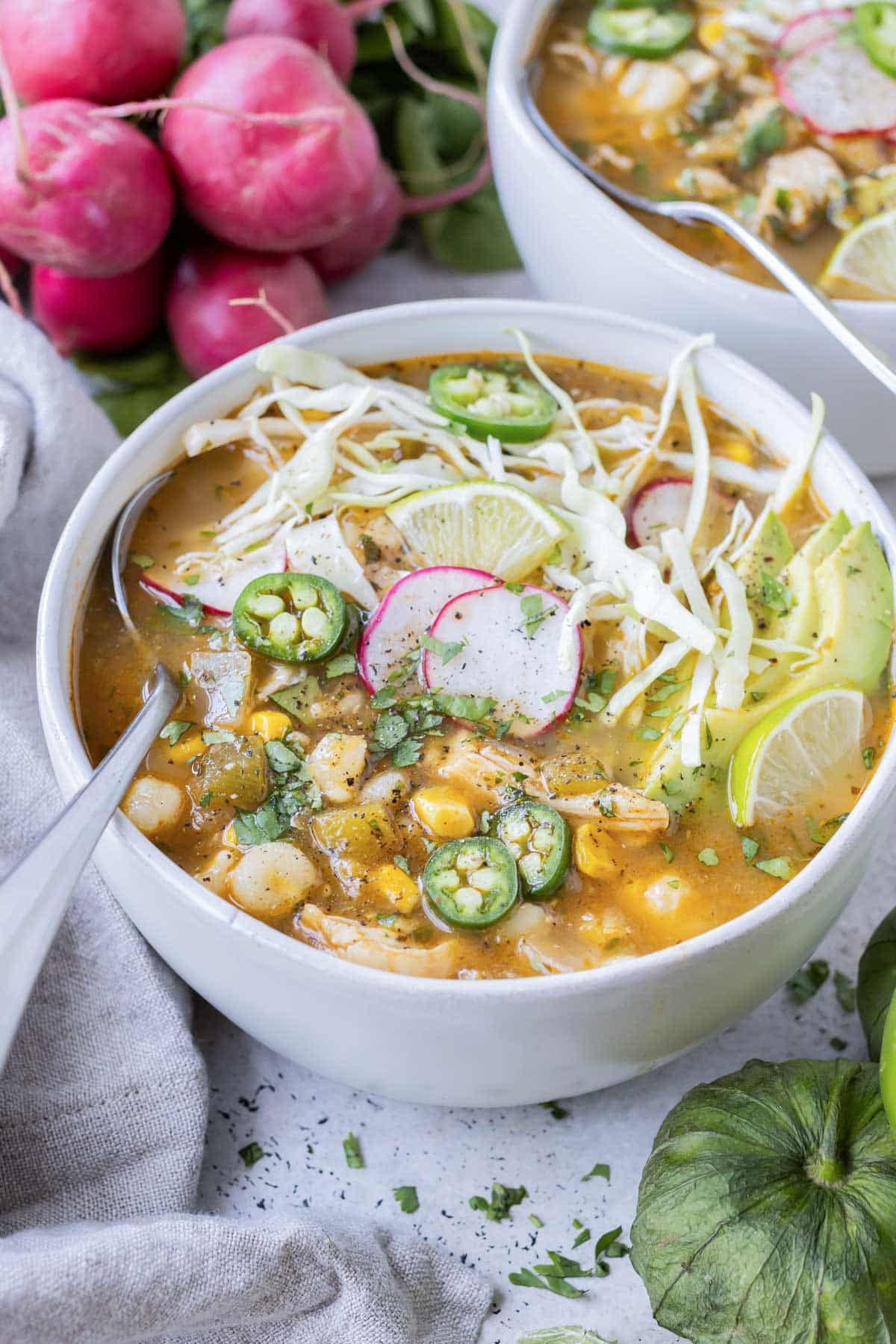 Two bowls are full of chicken pozole verde and served with fresh radish next to a napkin.