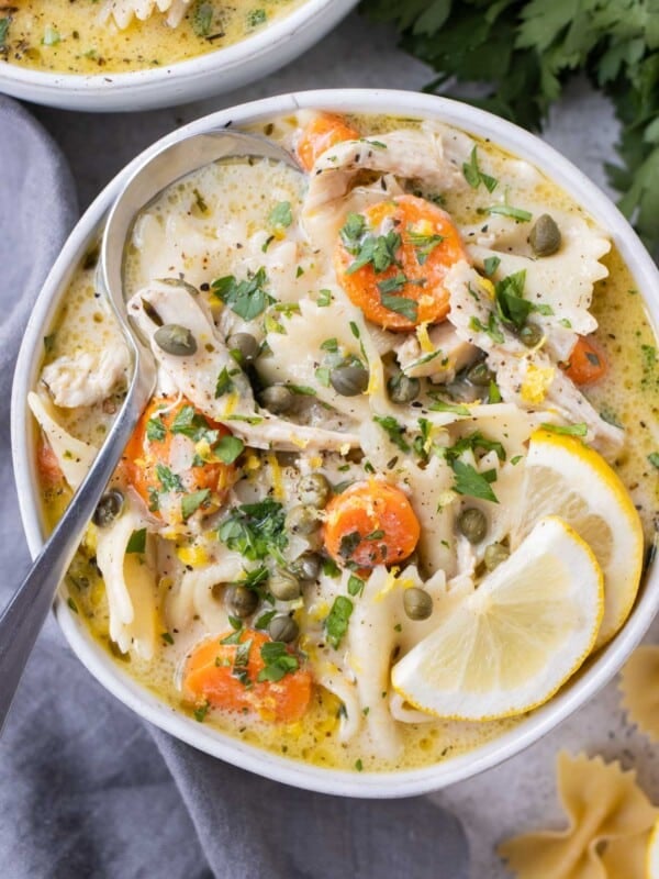 A bowl full of a creamy lemon chicken piccata soup with carrots, pasta, capers, and lemon slices.