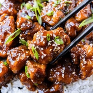 Chopsticks are picking up a piece of General Tso's chicken from a dish filled with white rice.