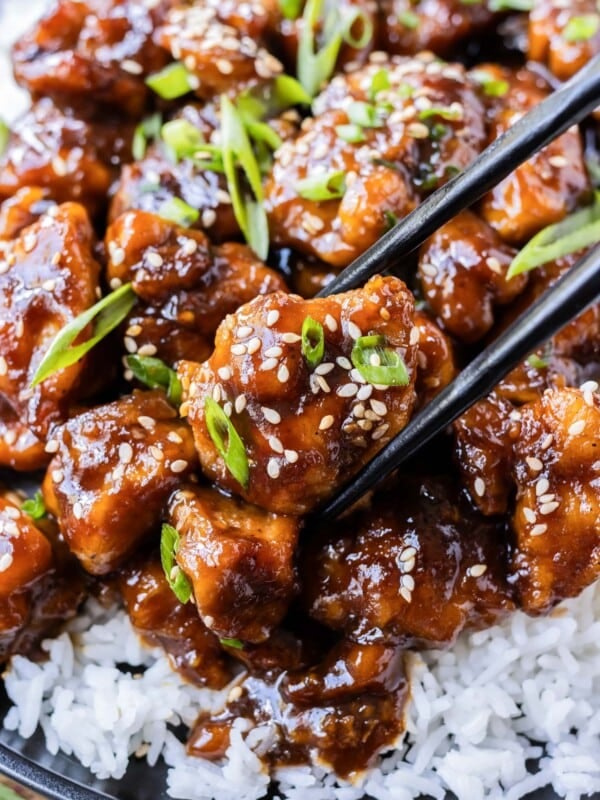 Chopsticks are picking up a piece of General Tso's chicken from a dish filled with white rice.
