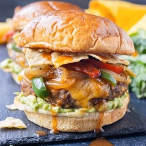 A beef enchilada burger with cheddar cheese, fajita veggies, and guacamole is served on a plate.