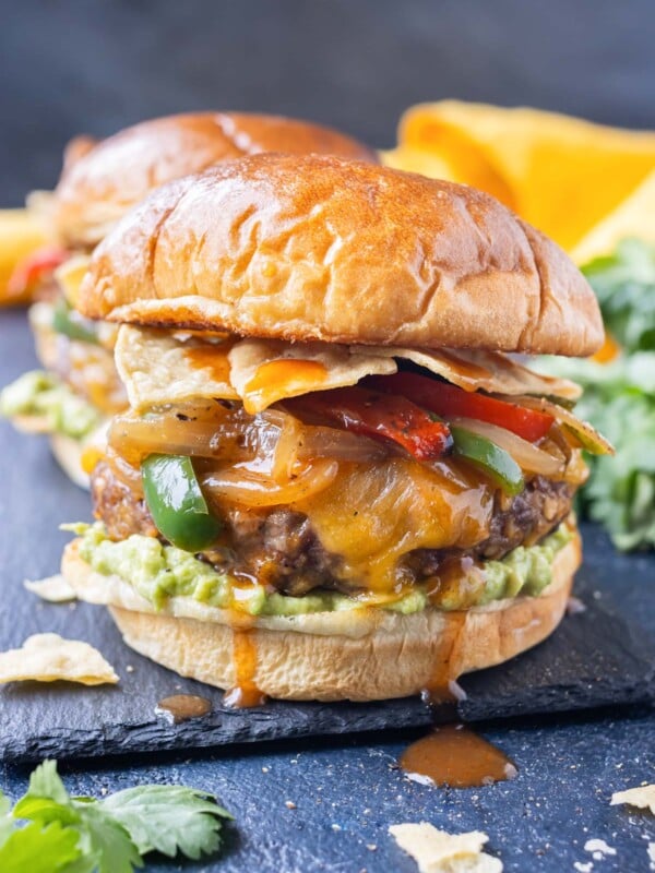 A beef enchilada burger with cheddar cheese, fajita veggies, and guacamole is served on a plate.