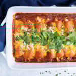 A shredded chicken enchiladas recipe with a homemade red sauce are in a baking dish with cheese and cilantro.