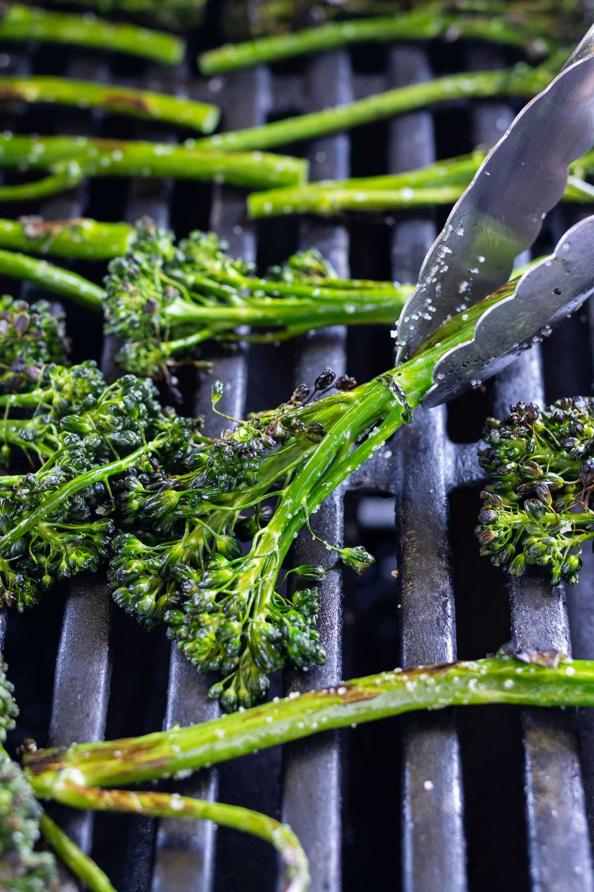 Tongs pick up a broccolini stalk from the grill.