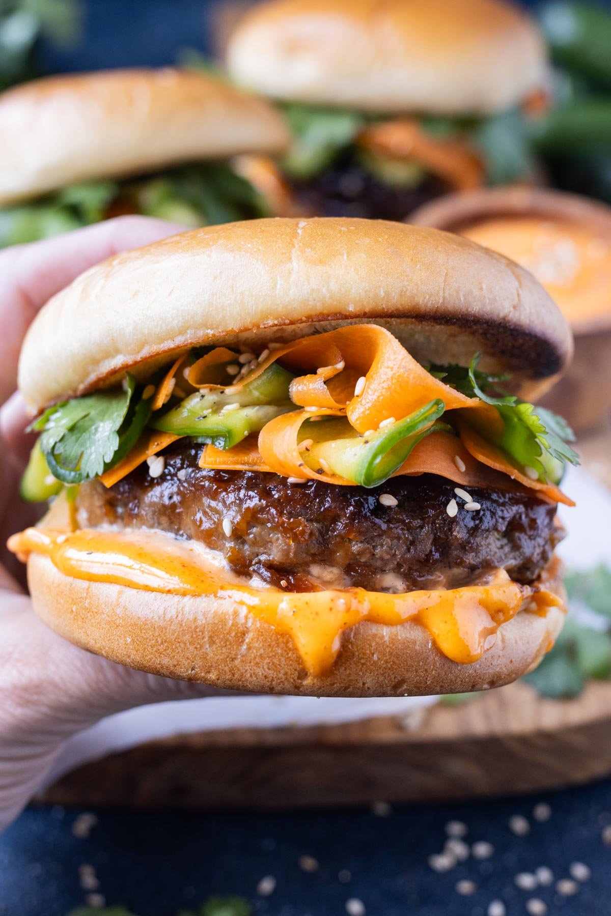 A hand is holding a Korean Beef Burger with gochujang mayo drizzling over the side.