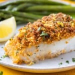 A fillet of Parmesan Crusted Cod is served on a plate with a lemon wedge and green beans.