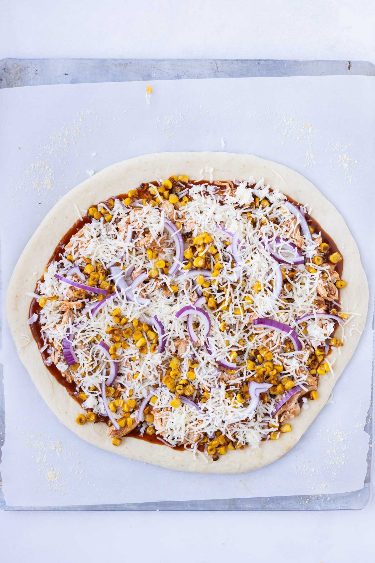 Cheese, chicken, red onion, and corn are added to the sauced pizza dough.
