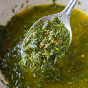 A spoon lifts up a homemade chimichurri sauce from a bowl with parsley, red pepper flakes, and oil.