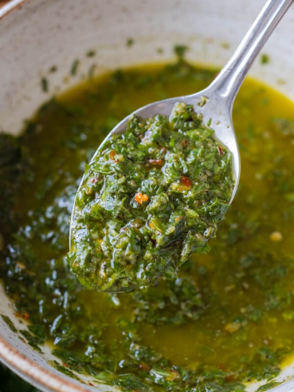 A spoon lifts up a homemade chimichurri sauce from a bowl with parsley, red pepper flakes, and oil.