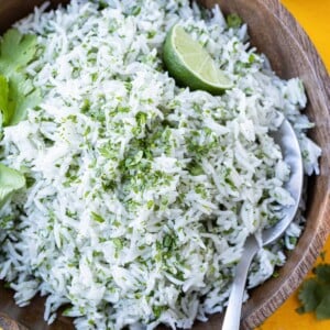 A wooden bowl is full of coconut milk cilantro lime rice with a lime wedge and a serving spoon.
