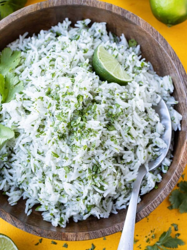 A wooden bowl is full of coconut milk cilantro lime rice with a lime wedge and a serving spoon.