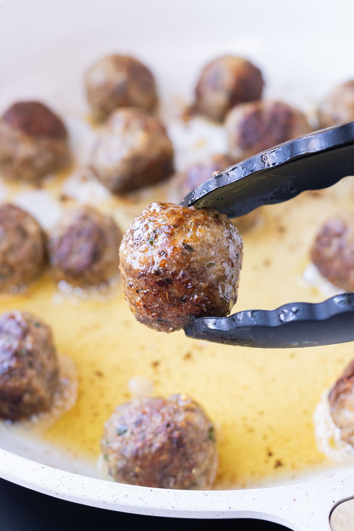 Tongs pick up a meatballs that has cooked in a skillet.