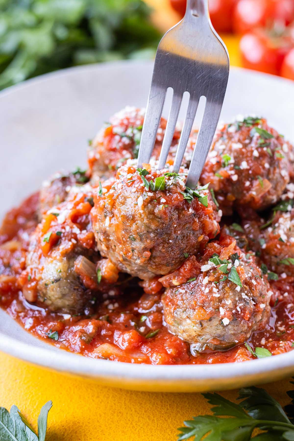 A fork picks up an Italian meatball from a white bowl with tomato sauce.