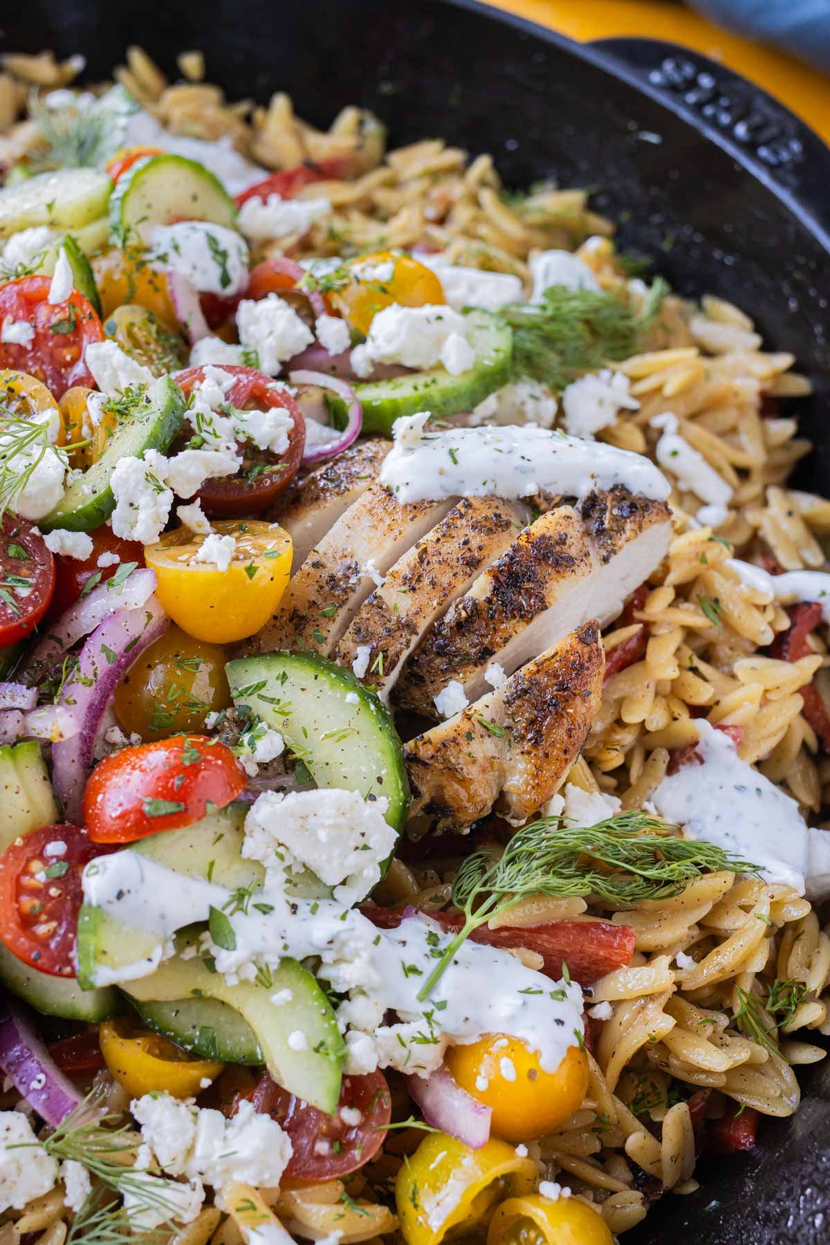 A skillet full of Greek chicken, creamy orzo pasta, cucumber tomato salad, and dill yogurt sauce is served for dinner.