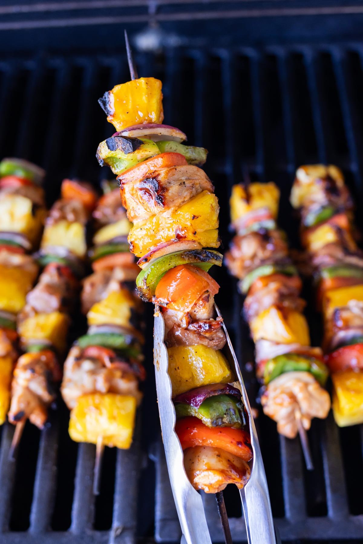 Tongs pick up a Hawaiian chicken kebab from a grill.