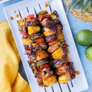 Hawaiian Chicken Kabobs are on a white plate with a soy marinade and pineapple next to them.