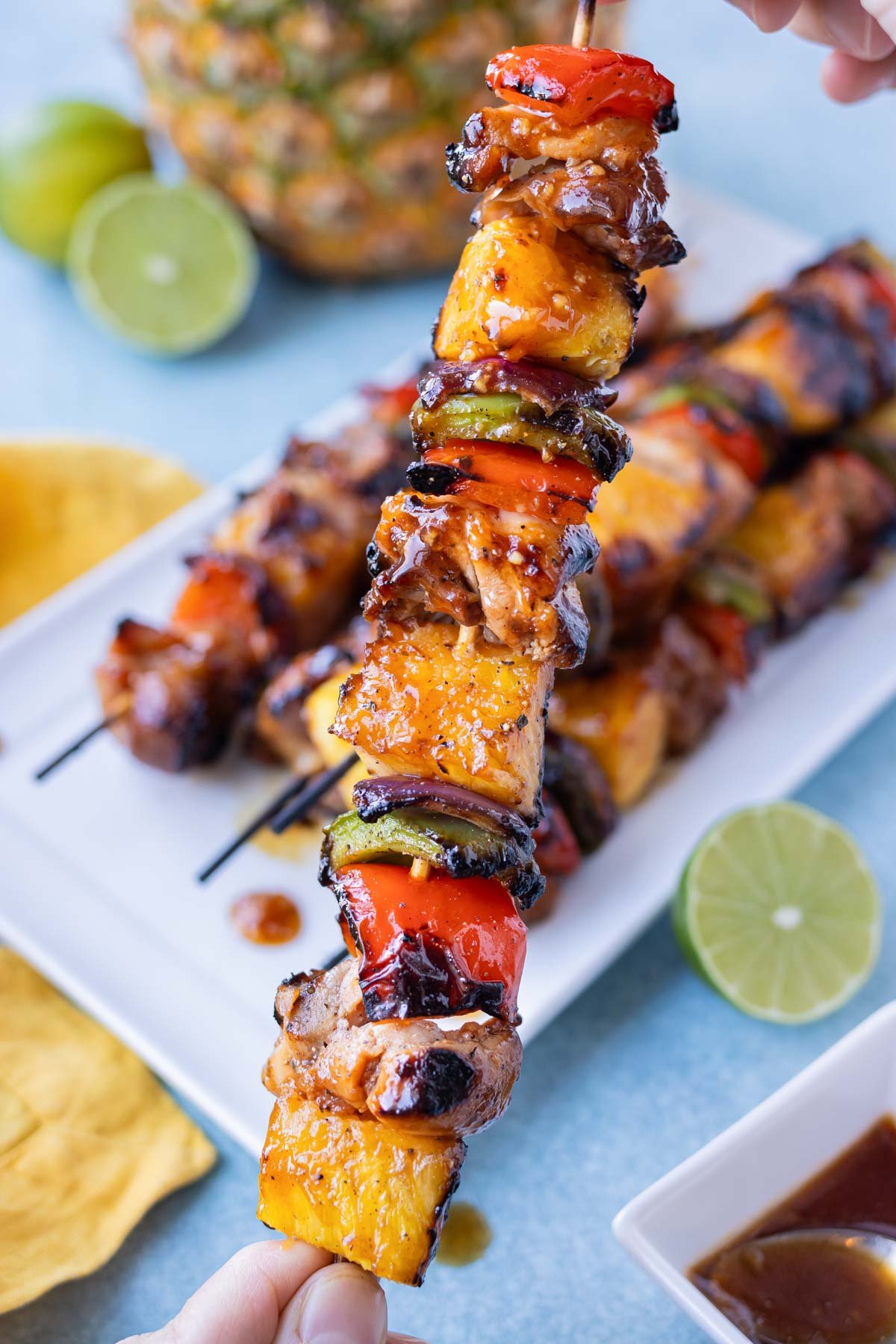A hand is holding a Hawaiian chicken kabob skewer with pineapples and bell peppers.