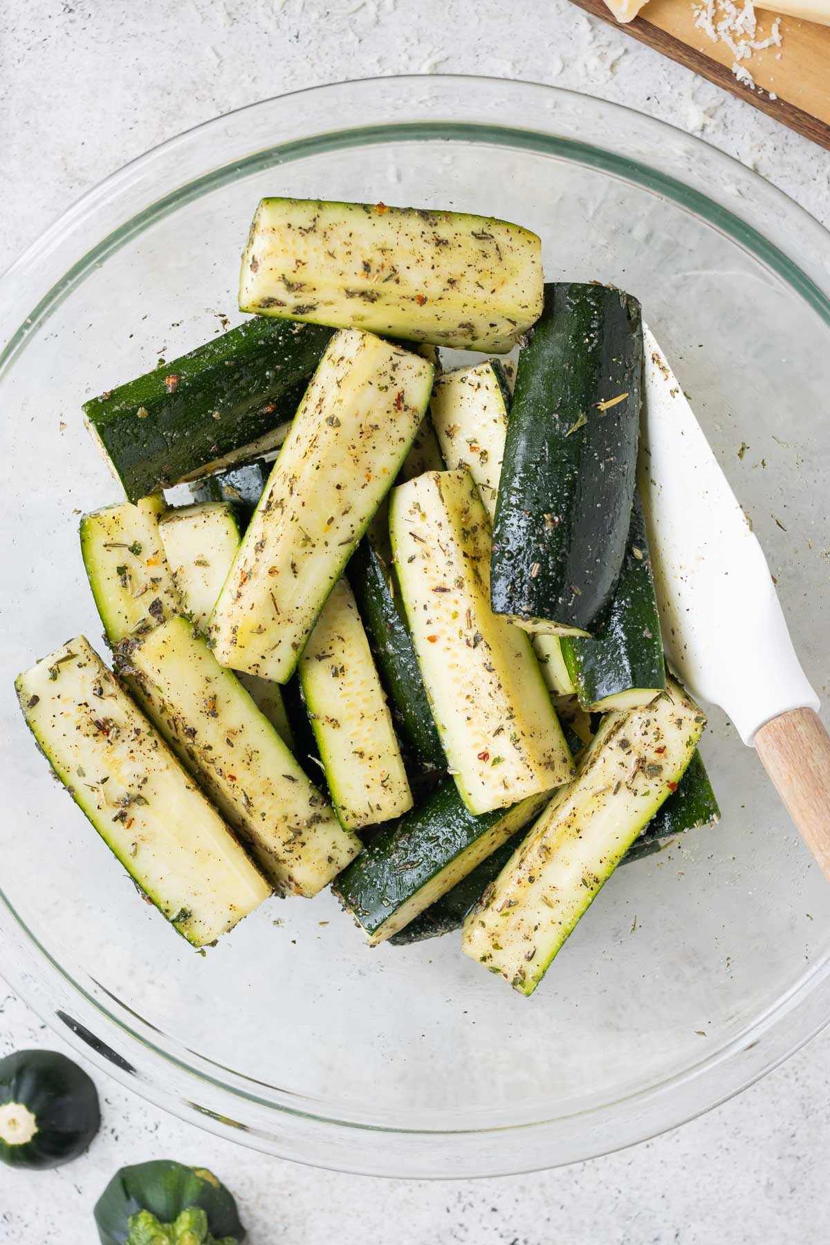 Sliced zucchini is tossed with oil and seasonings in a glass bowl.