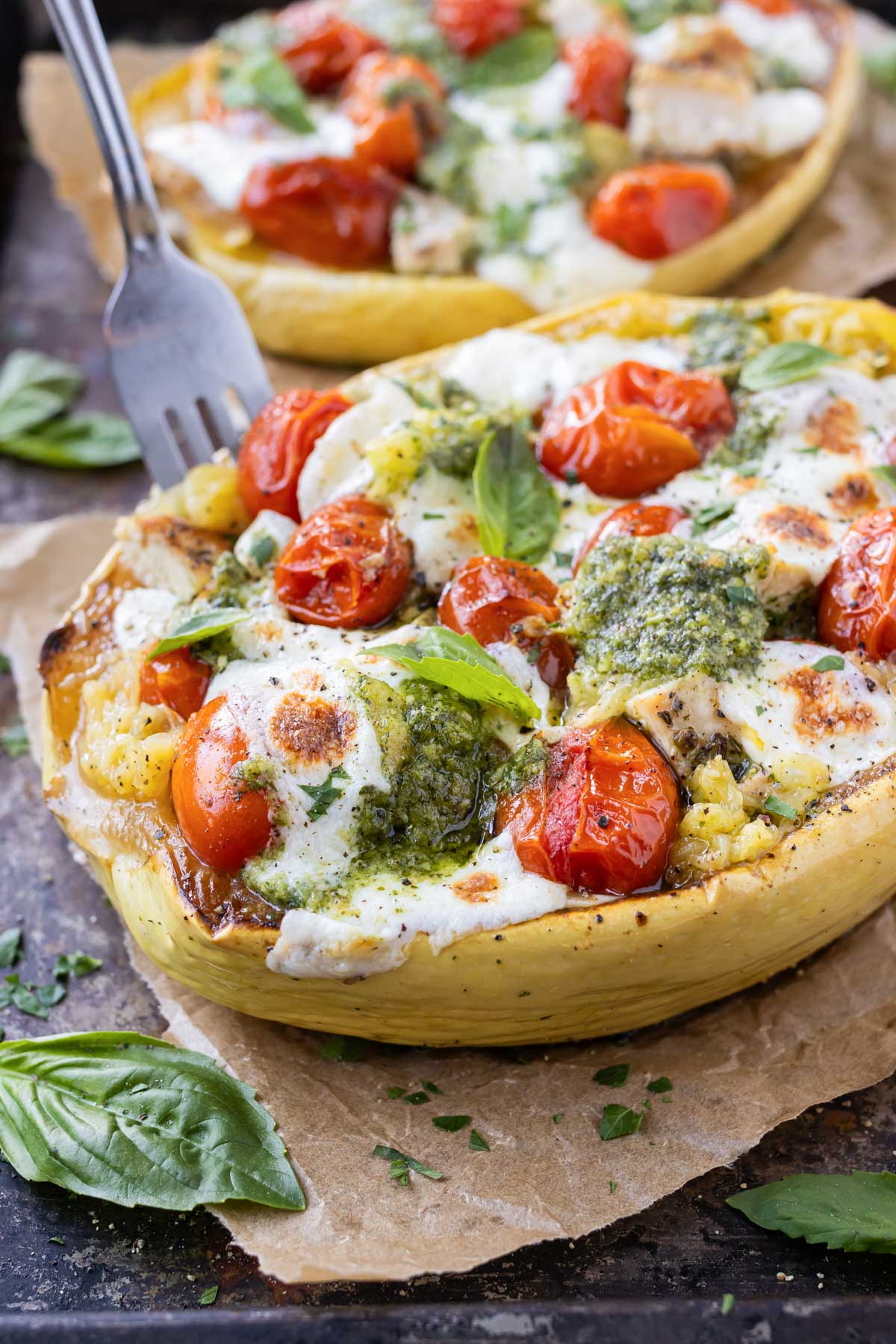 A fork digs into a spaghetti squash that is loaded with pesto sauce, chicken, cherry tomatoes, and mozzarella cheese.