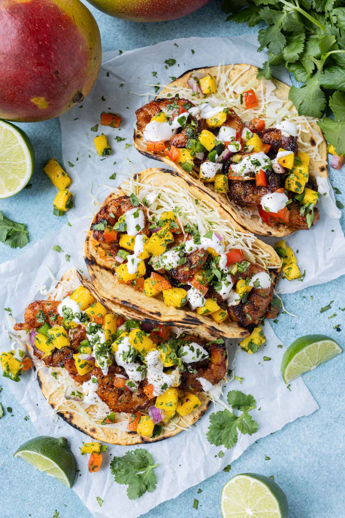 Three chili lime spiced shrimp tacos with a homemade mango salsa are next to limes and a mango.