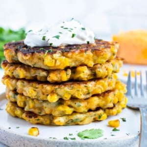 A stack of crispy corn fritters are on a white plate with sour cream on top.