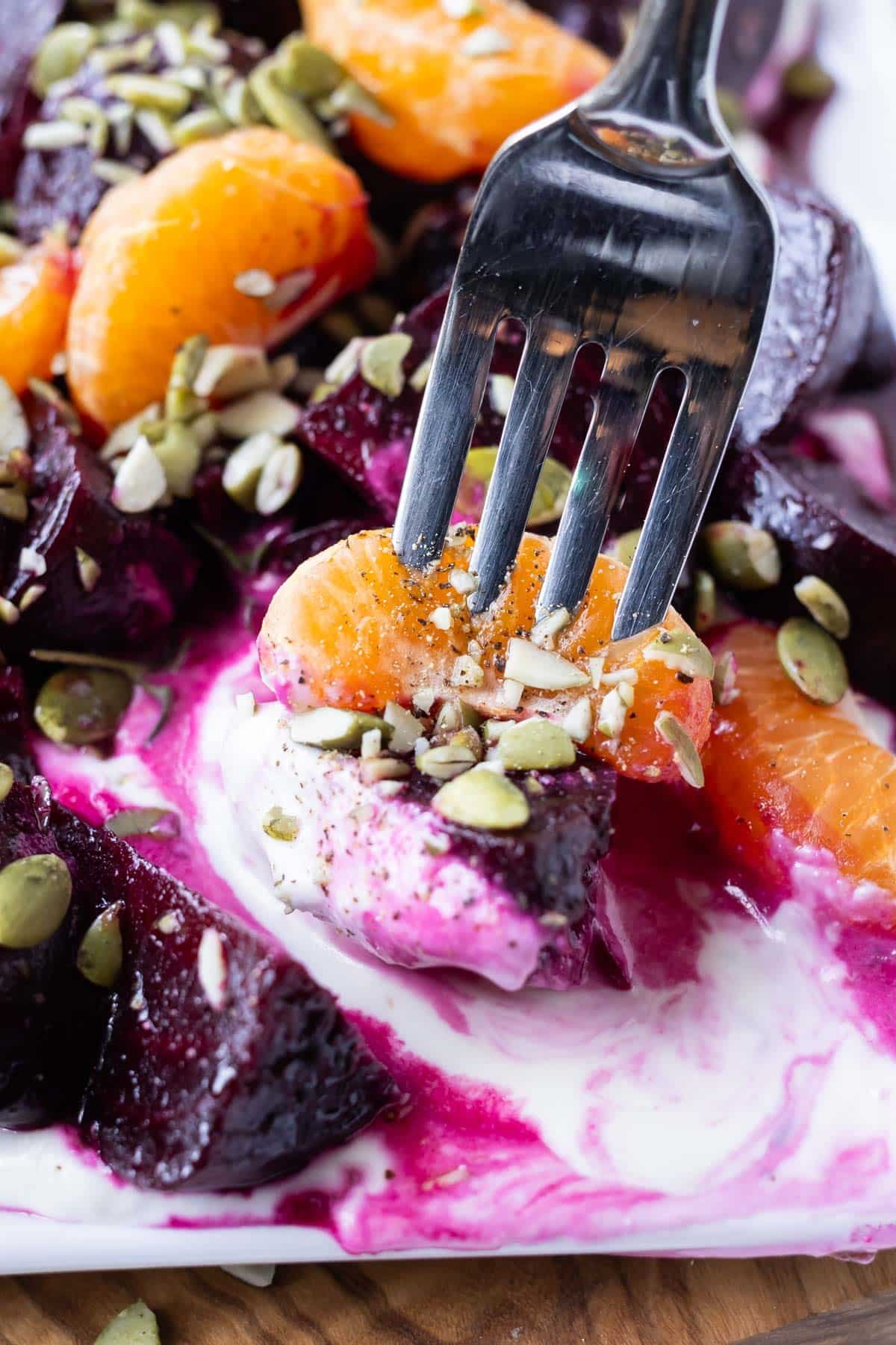 A fork picks up a bite of a roasted beet, an orange wedge, with roasted pumpkin seeds and whipped goat cheese.