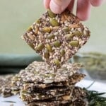 A hand is picking up a homemade seed cracker from a stack on a white plate.