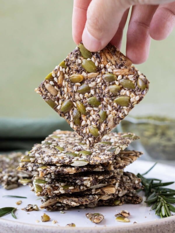 A hand is picking up a homemade seed cracker from a stack on a white plate.