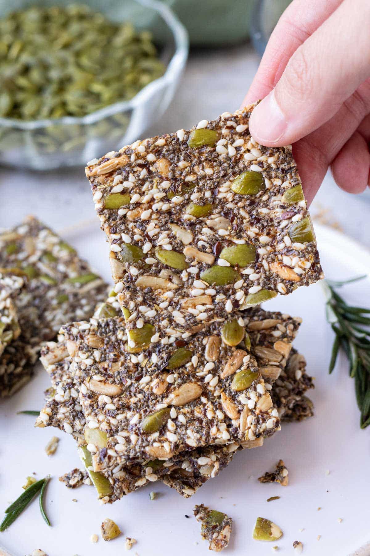 A hand is holding a homemade seed cracker from a stack of them on a white plate with pumpkin seeds and flax seeds.