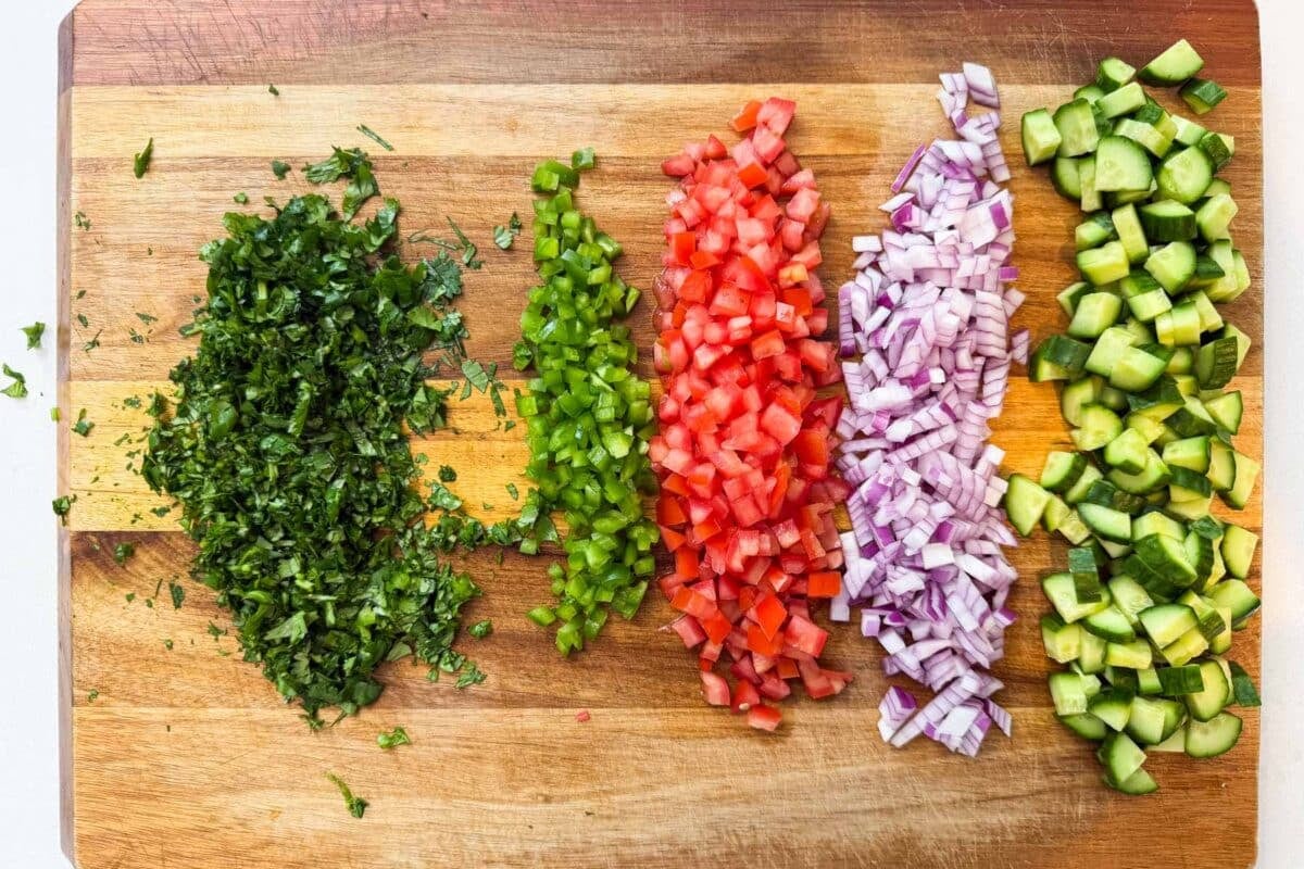 Diced cucumber, red onion, tomato, jalapeno and cilantro on a cutting board being prepped for a shrimp ceviche recipe.