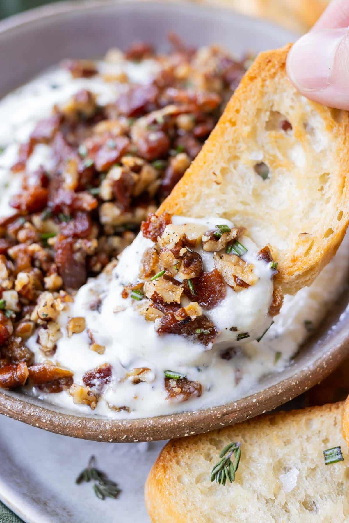 A hand is scooping up some fluffy whipped goat cheese with honey from a bowl that's topped with bacon bits.