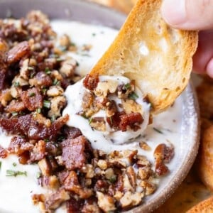 A crostini is being dipped into a bowl of whipped goat cheese with honey and a candied bacon and pecan topping.