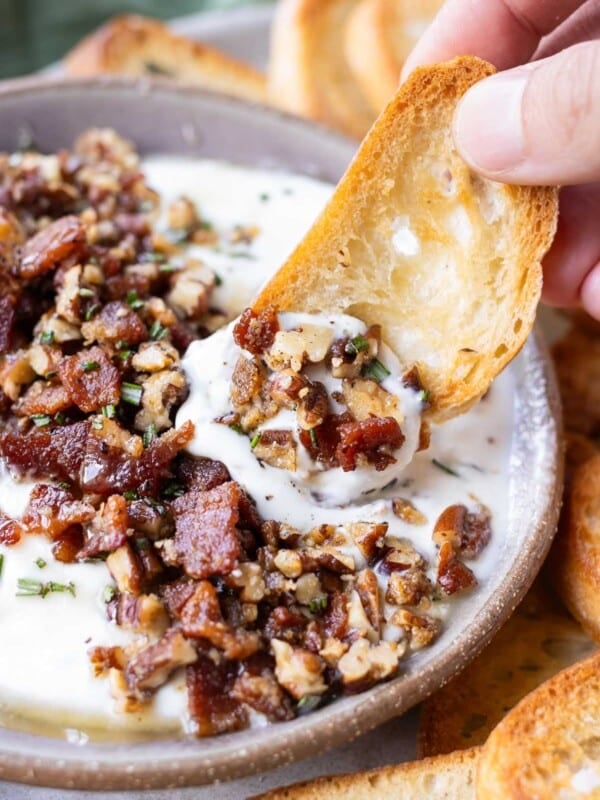 A crostini is being dipped into a bowl of whipped goat cheese with honey and a candied bacon and pecan topping.