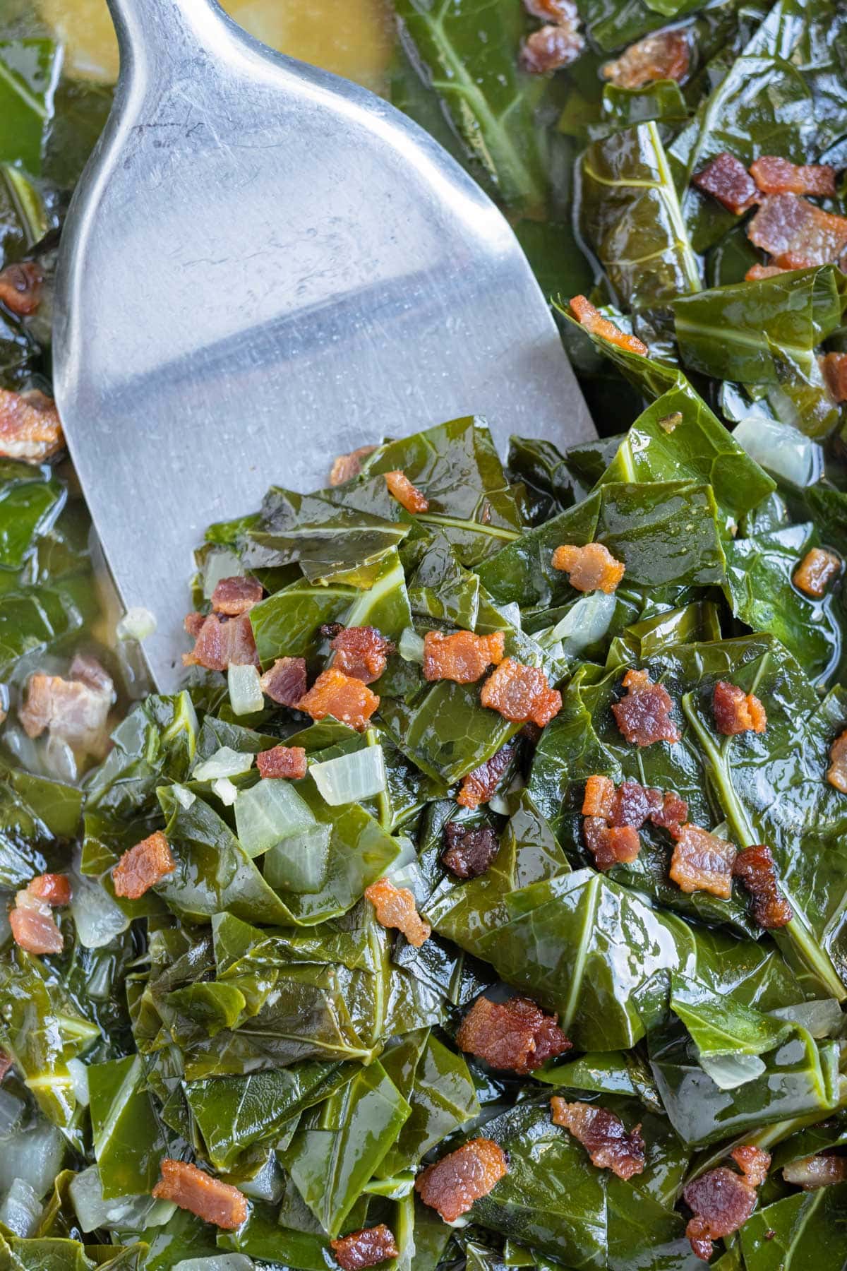 A spatula scoops up collard greens with bacon from a skillet for a New Year's dinner.