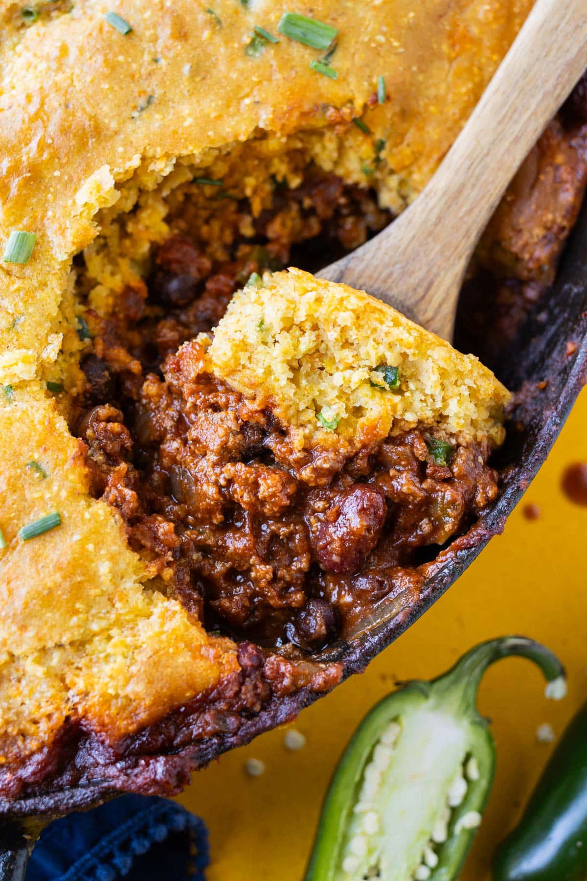 A wooden spoon scoops out a serving of a beef chili and cheddar jalapeño cornbread casserole from a skillet.