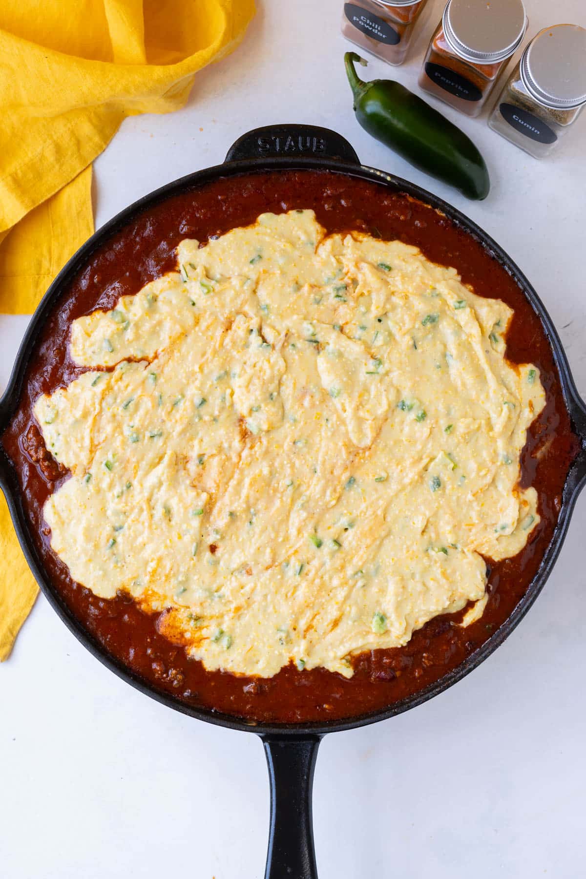 Prepared cornbread is spread across chili in a cast iron skillet.