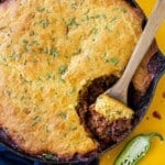 A cast-iron skillet is full of a chili cornbread casserole recipe with a wooden spoon scooping some out.