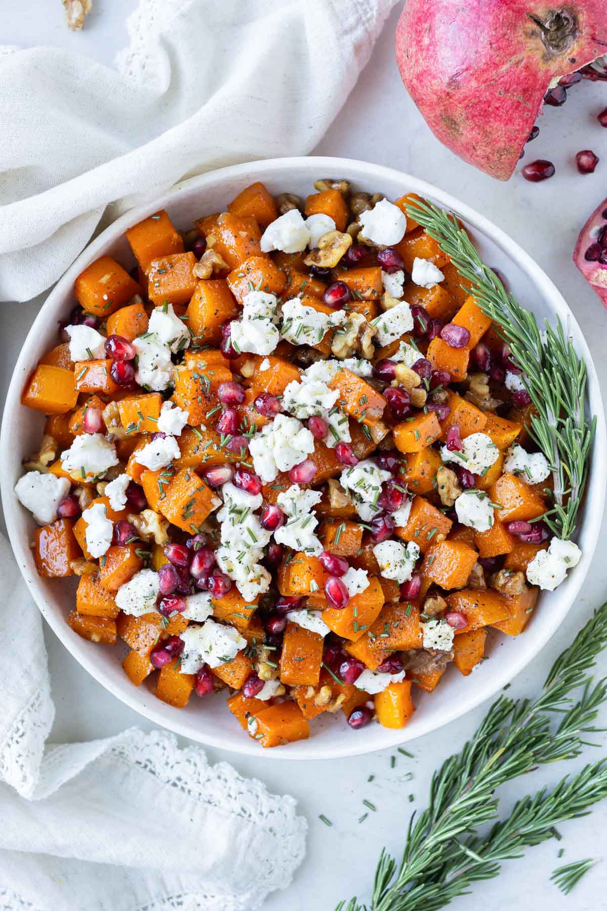 Honey roasted butternut squash cubes are in a white bowl with goat cheese, pomegranates, and walnuts.