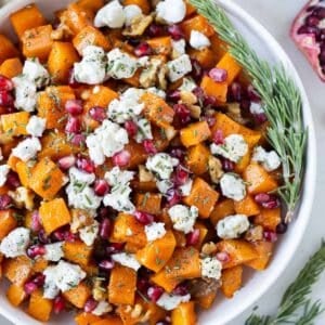 Roasted butternut squash cubes with goat cheese, pomegranates, walnuts, and rosemary are in a white serving bowl.