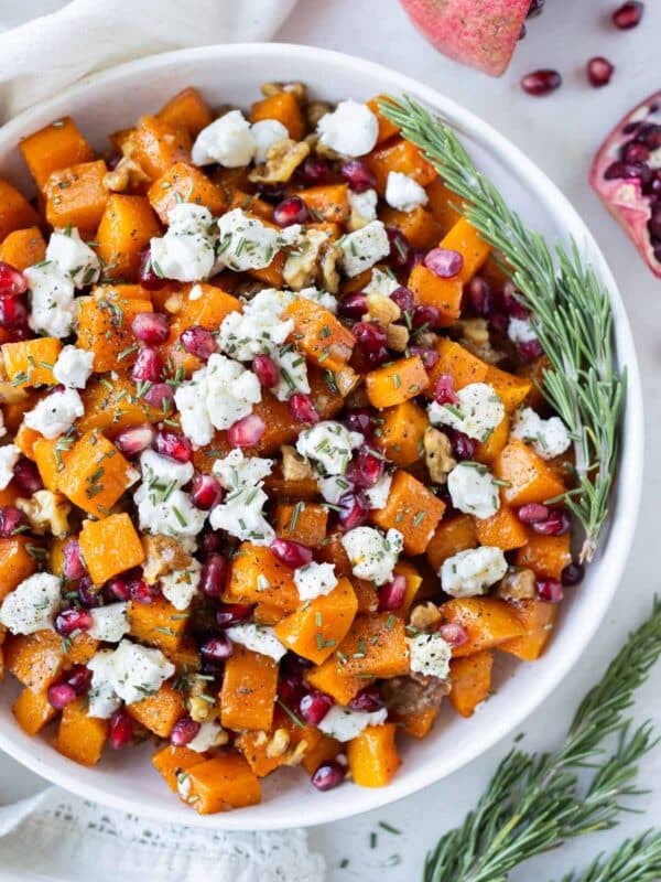 Roasted butternut squash cubes with goat cheese, pomegranates, walnuts, and rosemary are in a white serving bowl.