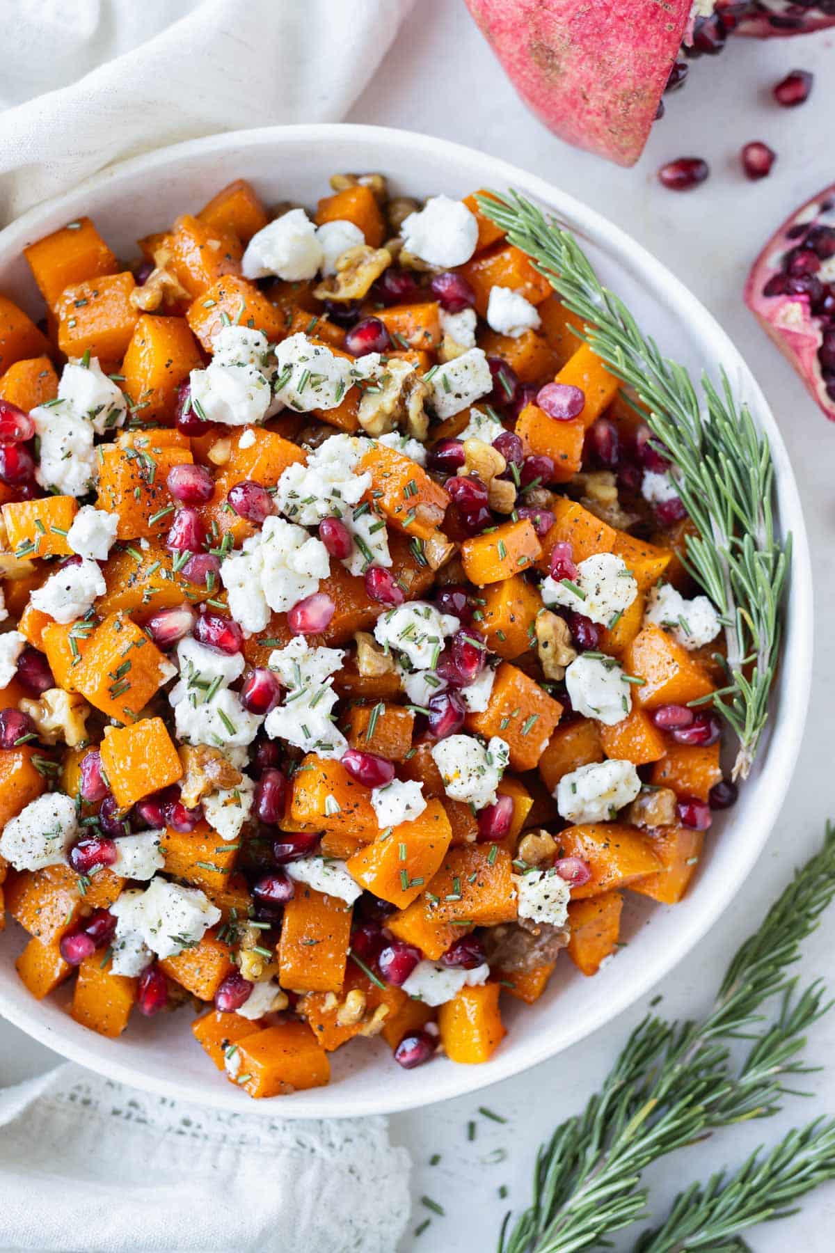 Roasted butternut squash cubes with goat cheese, pomegranates, walnuts, and rosemary are in a white serving bowl.