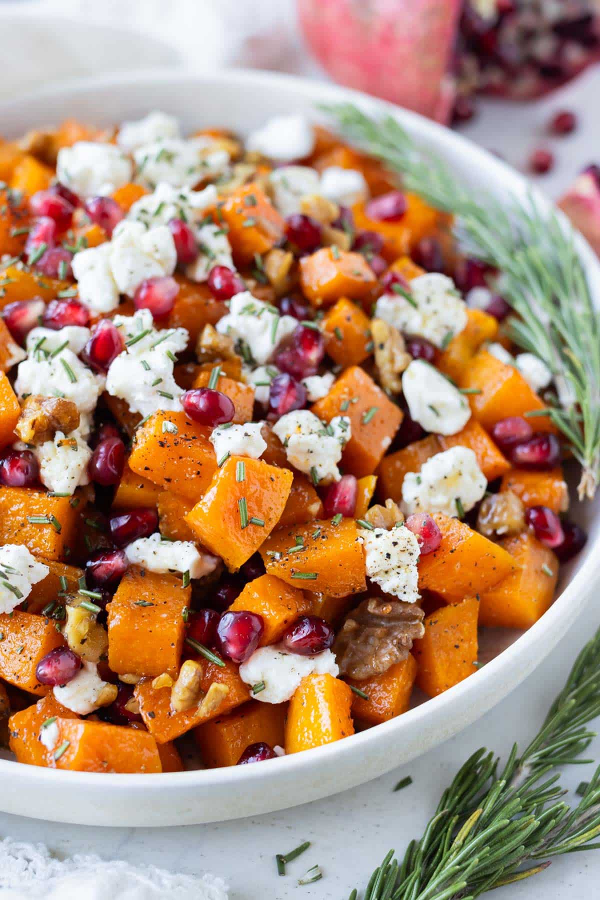A holiday side dish of baked butternut squash cubes with goat cheese, honey, pomegranates and walnuts is in a large bowl.